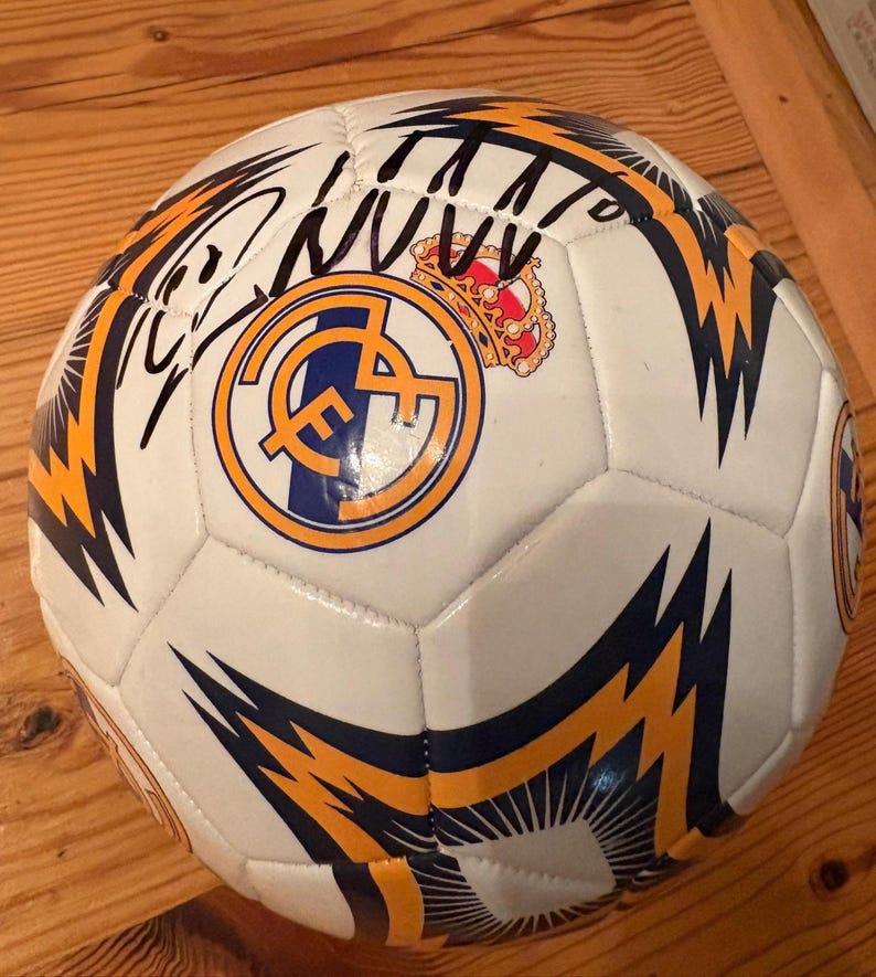 May include: A white soccer ball featuring the Real Madrid club crest in blue, gold, and red, accompanied by a black ink signature. The ball is decorated with dark blue and gold lightning bolt patterns across its multi-paneled surface.