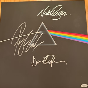 May include: A black vinyl record cover featuring the iconic prism and rainbow spectrum graphic from The Dark Side of the Moon. The cover displays three distinct white ink signatures and a small authentication sticker in the bottom right corner.