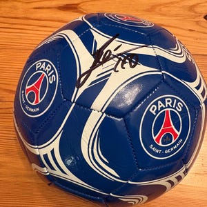 Pode incluir: Uma bola de futebol azul e branca com o logotipo do Paris Saint-Germain e uma assinatura. A bola tem um fundo azul com detalhes curvos brancos. O logotipo do Paris Saint-Germain está na bola em um design circular com a Torre Eiffel.