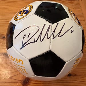May include: A white and black soccer ball with a signature and team logos. The ball has a classic pentagon and hexagon design. The team logos are gold and blue. The signature is in black ink.