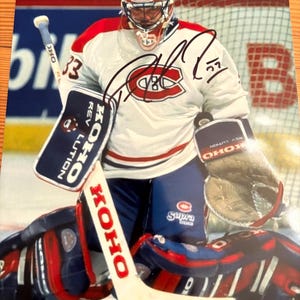 May include: A signed photograph of a hockey goalie in a white, red, and blue Montreal Canadiens uniform. The goalie wears a helmet and pads, holding a hockey stick. The image includes the number 33 and the KOHO logo.