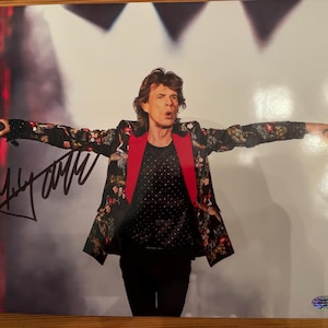 May include: A stage photograph of a man with arms outstretched. He wears a black jacket with floral patterns, a red collar, and a black shirt with white polka dots. The image is signed with a black marker.