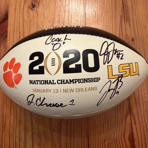 May include: A white and black football featuring the text 2020 NATIONAL CHAMPIONSHIP JANUARY 13 | NEW ORLEANS. The ball displays an orange paw print logo, the LSU logo, and multiple black ink signatures including Coach O and J. Chase 1.