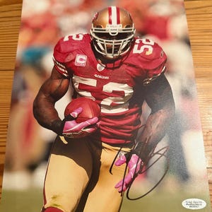 May include: A signed photograph of a football player in a red and gold uniform, holding a football. The player wears a helmet and pink gloves. The jersey has the number 52. The background is blurred, suggesting a game setting.