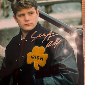 May include: A signed photograph featuring a man wearing a black jacket with a gold shamrock patch that reads "IRISH." The jacket is over a maroon shirt. The signature reads "Sey A. Rudy."