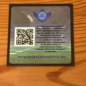 May include: A square authentication card from Total Sports Authentics featuring a blue logo with wings and a crown, a QR code, and text stating the item has been examined and deemed genuine. The background shows a green football field graphic.