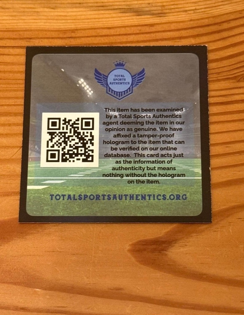 May include: A square authentication card from Total Sports Authentics featuring a blue logo with wings and a crown, a QR code, and text stating the item has been examined and deemed genuine. The background shows a green football field graphic.