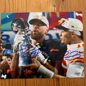 Puede incluir: Una fotografía en color que muestra a dos atletas con uniformes de los Kansas City Chiefs sosteniendo el trofeo Vince Lombardi. La imagen incluye autógrafos en tinta azul sobre el trofeo y la camiseta blanca, con una pegatina de Total Sports Authentics.
