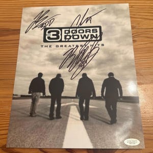 Puede incluir: Una impresión firmada del álbum "The Greatest Hits" de la banda 3 Doors Down. La imagen muestra a los miembros de la banda alejándose por una carretera, con el logotipo de la banda y firmas arriba. La impresión está sobre una superficie de madera.