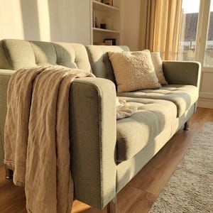 May include: A light green sofa with a beige throw blanket draped over the arm. A fluffy cream-colored pillow and a smaller beige pillow rest on the sofa. The sofa has silver legs and is in a well-lit room with a rug.