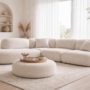 May include: A cream-colored sectional sofa with rounded edges and matching ottoman. The sofa has several throw pillows and sits on a patterned rug. A wooden side table with a lamp and a decorative shelf are also visible.