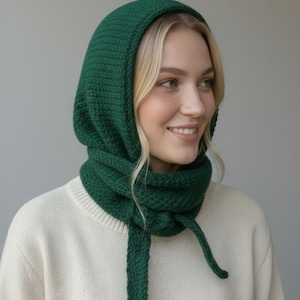 Op de afbeelding: Een gebreide groene capuchon- en sjaalset. De capuchon bedekt het hoofd en de nek, met een sjaal die kan worden vastgebonden. De set is gemaakt van een getextureerd, donkergroen garen. De persoon draagt een crèmekleurige trui.