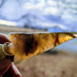 Może przedstawiać: Przezroczysty, bursztynowy wisiorek w kształcie grota strzały z ciemnobrązowymi wtrąceniami. Grot strzały jest przymocowany do małego, zaokrąglonego kawałka ze złotym pierścieniem. Tło jest rozmyte.