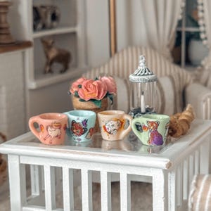May include: Four miniature ceramic mugs with animal designs sit on a white table. The mugs are pink, light blue, tan, and green. Each mug features a different animal illustration: a unicorn, a leopard, a dog, and a leopard. A small bouquet of pink roses is in the background.
