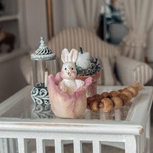 May include: A miniature Easter scene featuring a white bunny figurine nestled in a pink and white flower-shaped container. A decorative lantern and a small potted plant with a candle are also present. A string of brown decorative balls rests on a white table.