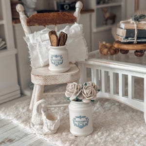 May include: Miniature white ceramic jars with blue floral designs and text. One jar reads "BAGUETTES" and holds wooden utensils. Another jar reads "USTENSILE" and holds fabric roses. The jars are part of a dollhouse scene.