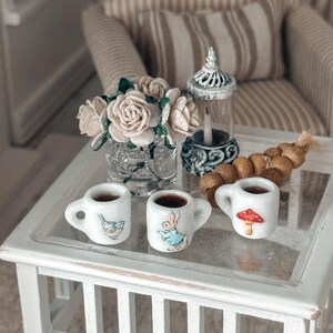 May include: A miniature tea set with three white mugs, each with a different illustration. One mug has a bird, another a rabbit, and the third a red mushroom. A small vase of roses, a candle holder, and a decorative item are also on the table.