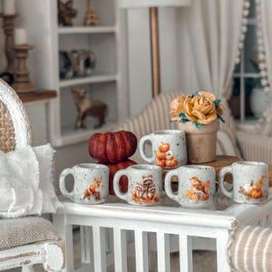 May include: A collection of miniature ceramic mugs with autumn-themed designs. Each mug features a different illustration, including pumpkins, a fox, a raccoon, and a bird. The mugs are displayed on a white table, alongside a small pumpkin and a pot of flowers.