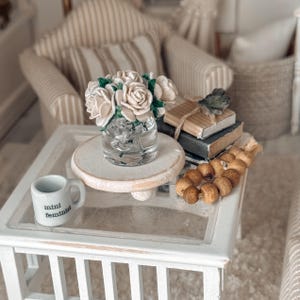 May include: A miniature white table with a glass top displays a small bouquet of cream-colored roses in a clear glass vase. A tiny mug with the words "mini feminist" is also on the table, along with a stack of books and decorative wooden beads.