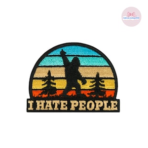 May include: Embroidered patch featuring a black silhouette of Bigfoot with an arm raised, set against a sunset of blue, orange, and yellow stripes. The text "I HATE PEOPLE" is displayed in bold, white letters.
