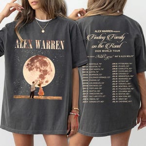 以下が含まれることがあります： チャコールグレーのTシャツで、月夜の風景の中で踊る二人の人物の大きなグラフィックが特徴です。前面には「ALEX WARREN」の文字があり、背面には「Finding Family on the Road 2026 World Tour」の日程と開催地が記載されています。