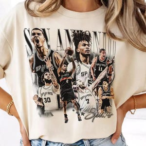 May include: Cream-colored t-shirt featuring a graphic of San Antonio Spurs basketball players. The design includes the team name and logo, with players in black and white uniforms. The shirt has a vintage, distressed look.