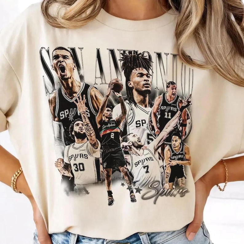 May include: Cream-colored t-shirt featuring a graphic of San Antonio Spurs basketball players. The design includes the team name and logo, with players in black and white uniforms. The shirt has a vintage, distressed look.