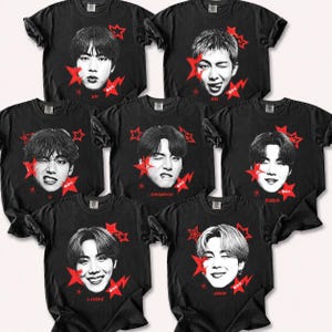 Op de afbeelding: Een collectie van zeven zwarte t-shirts met korte mouwen, elk voorzien van een contrastrijk zwart-wit portret van een ander persoon. Elk shirt bevat rode stergraphics en de naam van de persoon onder hun portret gedrukt.