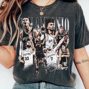 May include: Dark gray t-shirt featuring a graphic of San Antonio Spurs basketball players. The design includes the team name and the word "Spurs" in a stylized font. The players are in black and white uniforms.