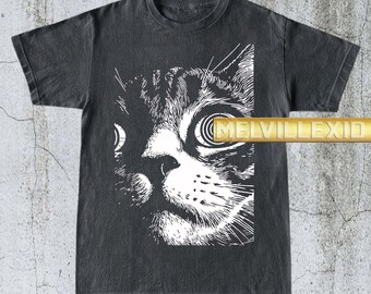 May include: Charcoal grey t-shirt with a white graphic of a cat's face. The design highlights the cat's eye and nose. The text "MELVILLEXIO" is printed on the shirt.