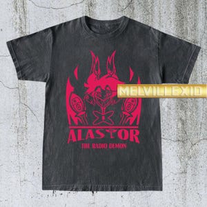 May include: Dark gray t-shirt featuring a red graphic of a cartoon character with the text "ALASTOR THE RADIO DEMON". The shirt has a vintage, distressed look.