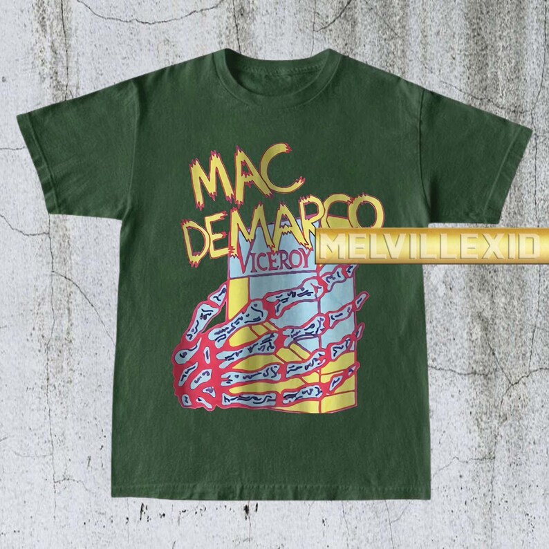 以下が含まれることがあります： 黄色と赤で「MAC DEMARCO VICEROY」の文字が入った濃い緑色のTシャツ。 骸骨の手のグラフィックが、青と黄色のデザインの箱を持っています。 Tシャツはクルーネックで半袖です。