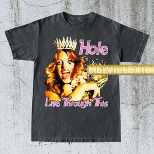 Puede incluir: Camiseta gris oscuro con un gráfico vintage. El diseño incluye una mujer con corona, la palabra "Hole" en rosa y la frase "Live Through This". La camiseta tiene un aspecto desgastado.