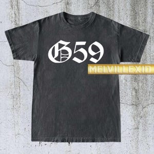 Puede incluir: Camiseta gris oscuro con el número "659" en blanco, con fuente gótica. La camiseta tiene un aspecto vintage. El texto "MELVILLEXID" está impreso en dorado en la parte inferior derecha.