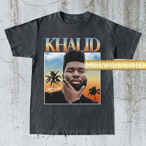 May include: Dark gray t-shirt featuring a graphic of the singer Khalid. The design includes the name "KHALID" in a gradient font, a portrait of the singer, palm trees, and the words "FREE SPIRIT".