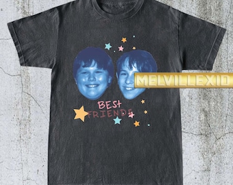May include: Dark gray t-shirt featuring two smiling faces with blue skin tones and the words "BEST FRIENDS" in white. Colorful stars and a starburst accent the design. The shirt has a vintage, worn look.