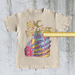 以下が含まれることがあります： ベージュ色のTシャツ。黄色と赤色の文字で「MAC DEMARCO VICEROY」と書かれています。グラフィックには、カラフルな長方形のデザインを握る骸骨の手が含まれています。シャツには「MELVILLEXID」の文字も入っています。