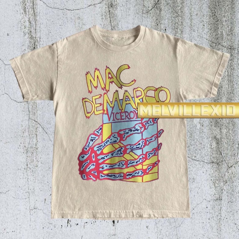 以下が含まれることがあります： ベージュ色のTシャツ。黄色と赤色の文字で「MAC DEMARCO VICEROY」と書かれています。グラフィックには、カラフルな長方形のデザインを握る骸骨の手が含まれています。シャツには「MELVILLEXID」の文字も入っています。