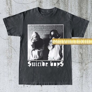 Peut inclure: T-shirt gris foncé avec une image en noir et blanc de deux personnes en sweat à capuche et le texte "Suicide Boys" en police gothique. L'image est placée sur un fond de mur de briques.