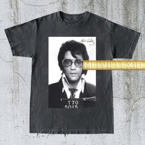 May include: Dark gray t-shirt featuring a black and white image of Elvis Presley in a mugshot. The image includes Elvis's signature and the numbers T70 5015. The shirt has a vintage, worn look.