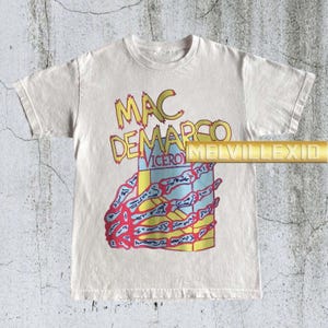 以下が含まれることがあります： オフホワイトのTシャツ。黄色と赤で「MAC DEMARCO VICEROY」の文字がプリントされています。グラフィックには、骸骨の手と箱のカラフルなデザインが含まれています。このシャツはクルーネックです。