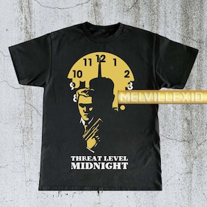 May include: Black t-shirt featuring a yellow clock design with a silhouette of a man holding a gun. The text "THREAT LEVEL MIDNIGHT" is printed in white below the graphic. The shirt is a classic crew neck style.