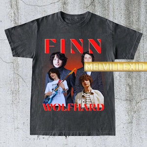 May include: Dark gray t-shirt featuring a vintage-style graphic of Finn Wolfhard with the text "FINN WOLFHARD" in red. The design includes multiple images of the actor. The shirt has a distressed look.