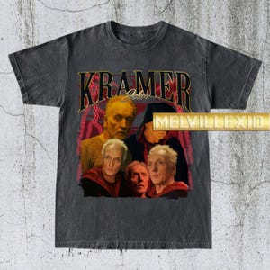 Könnte beinhalten: Dunkelgraues T-Shirt mit einer Grafik von John Kramer aus der Saw-Filmreihe. Das Design enthält den Namen "KRAMER" in goldfarbenen Buchstaben, mit einer Collage von Bildern der Figur. Das T-Shirt hat einen Vintage-Look.