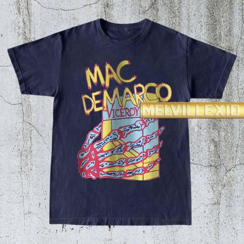 以下が含まれることがあります： 紺色のTシャツ。黄色と赤で「MAC DEMARCO」の文字、骸骨の手が箱を持っているグラフィック。 「VICEROY」の文字も見える。 ヴィンテージ感のあるTシャツ。