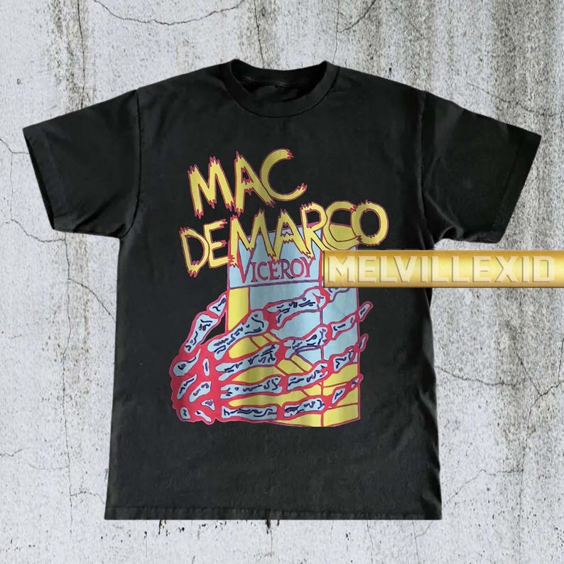 以下が含まれることがあります： 黄色とオレンジ色の「MAC DEMARCO」、青色の「VICEROY」の文字が入った黒いTシャツ。 骸骨の手のグラフィックがタバコの箱を持っています。 ヴィンテージ感のあるTシャツです。