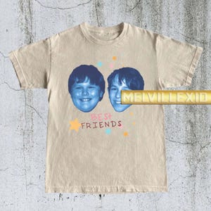 May include: Beige t-shirt featuring two blue-toned faces and the text "BEST FRIENDS" in pink and orange. The design includes star accents. The shirt has short sleeves and a crew neck.