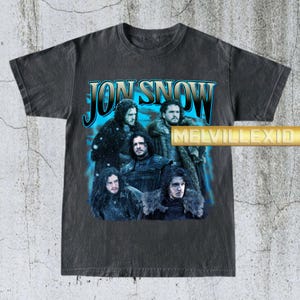 May include: Dark gray t-shirt featuring a graphic of Jon Snow from the television series. The design includes the text "JON SNOW" in blue and silver, with multiple images of the character. The shirt has a vintage, distressed look.