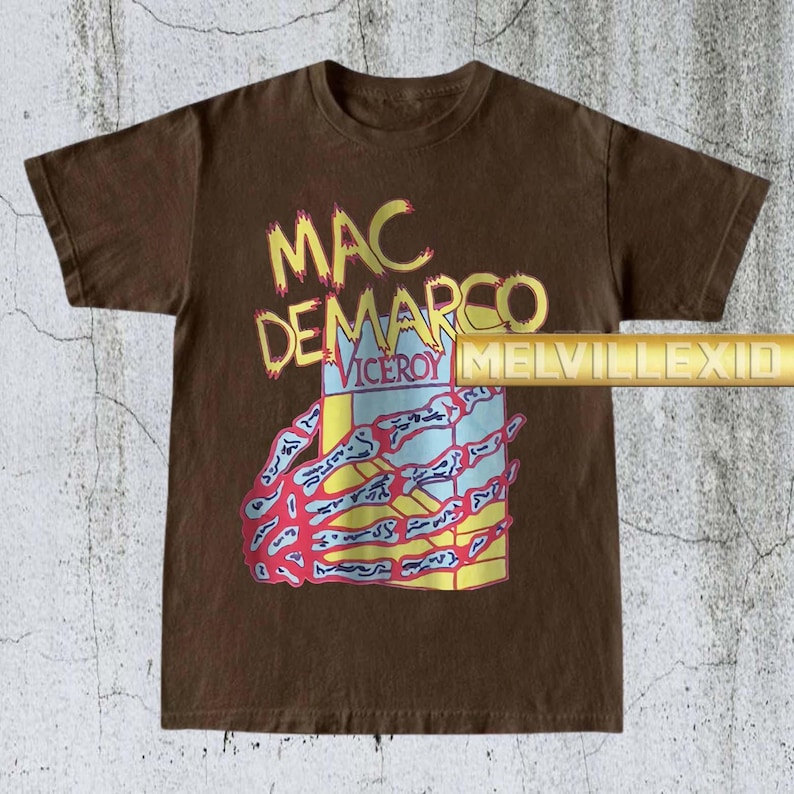 以下が含まれることがあります： 茶色のTシャツ。黄色で「MAC DEMARCO」と「VICEROY」の文字。青と赤のデザインの箱を持つ骸骨の手のグラフィック。ヴィンテージ感のあるTシャツ。