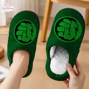 Pode incluir: Um par de chinelos de pelúcia verde-escuro com um emblema circular preto com um gráfico de punho verde na parte superior. O interior dos chinelos é forrado com um material branco texturizado semelhante a lã, com um aspeto macio e felpudo.